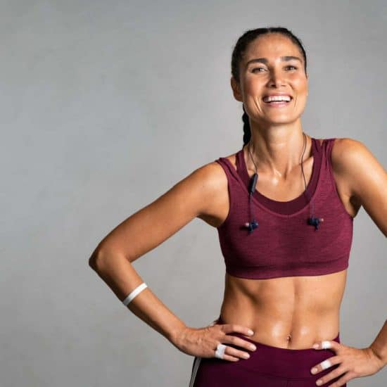 Portrait of beautiful fitness woman smiling and looking at camera isolated on grey background. Laughing mid woman in sportswear wearing wireless headphones and relaxing after training at gym. Happy fit girl excited about workout on gray wall with copy