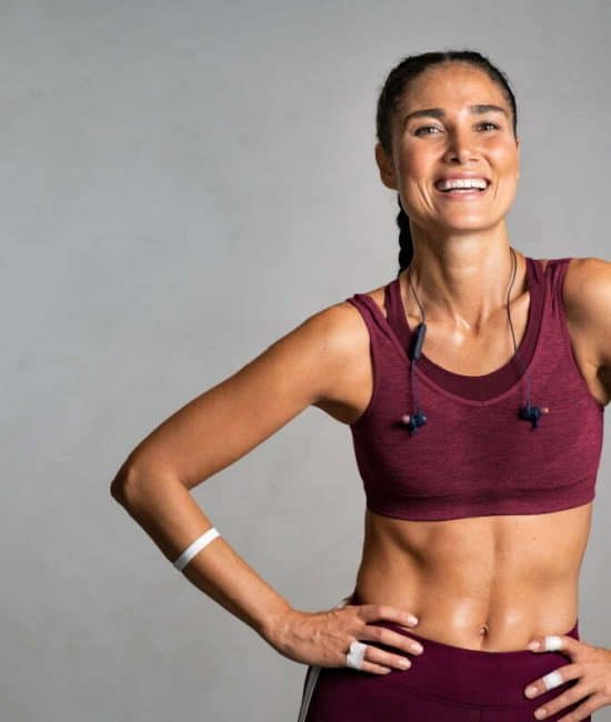 Portrait of beautiful fitness woman smiling and looking at camera isolated on grey background. Laughing mid woman in sportswear wearing wireless headphones and relaxing after training at gym. Happy fit girl excited about workout on gray wall with copy