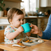 A baby drinks from a small blue cup at a high chair during a meal, with a woman smiling nearby.