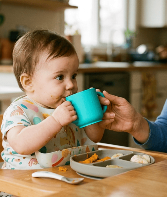 A baby drinks from a small blue cup at a high chair during a meal, with a woman smiling nearby.