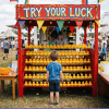 A young boy stands facing a bright red carnival game booth labeled TRY YOUR LUCK filled with rows of yellow rubber ducks, surrounded by a crowd at an outdoor fair under a partly cloudy sky.