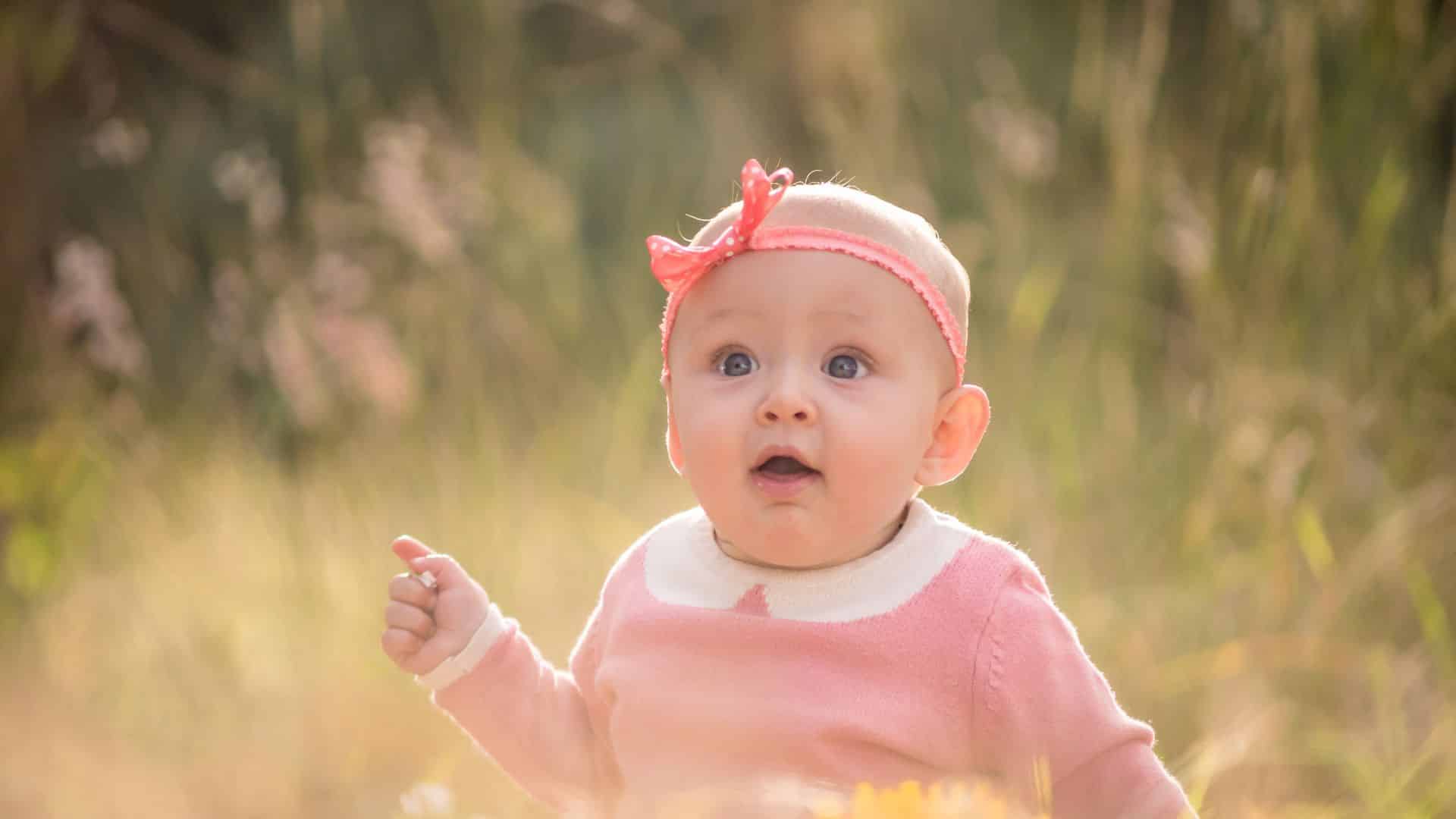 Baby girl with pink headband sitting in a sunlit field, wide-eyed and smiling, soft golden background capturing a joyful outdoor moment