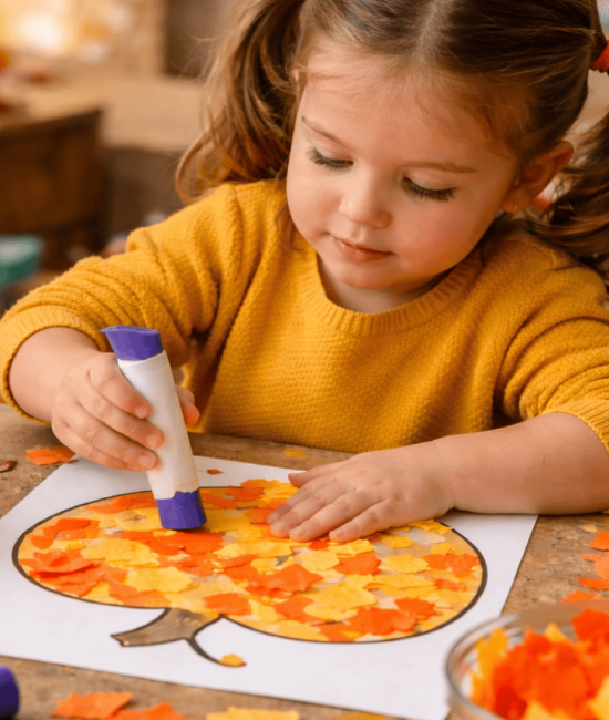 Child making a torn paper pumpkin mosaic craft, gluing orange and yellow tissue pieces onto a pumpkin outline at a fall-themed craft table.