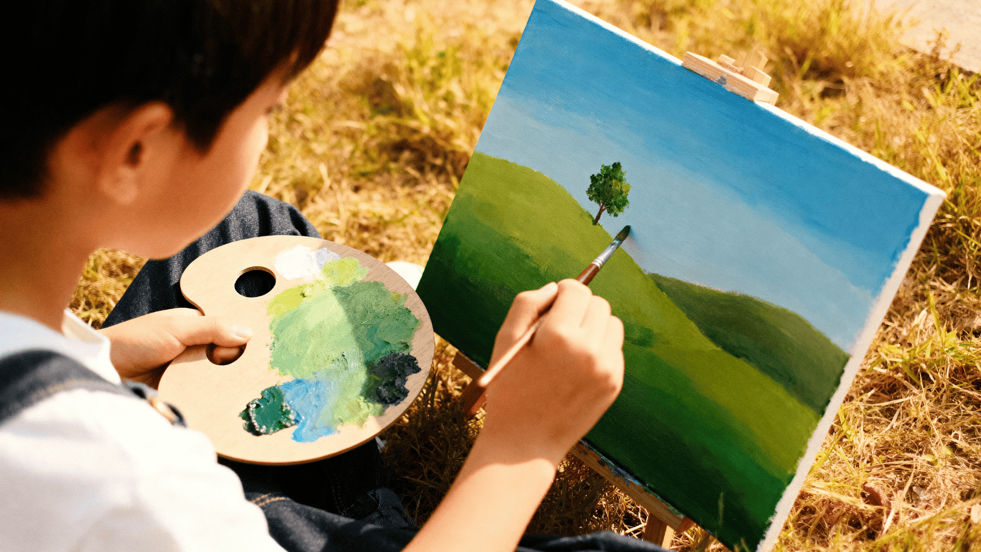 Child painting a simple landscape on canvas outdoors, holding a palette of green and blue paints, focusing on a lone tree on a grassy hill.