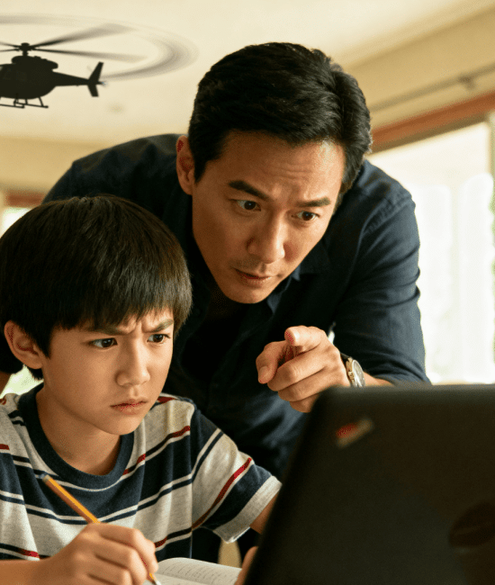 Father leans over child doing homework on laptop, pointing at screen while a toy helicopter flies overhead in a bright living room