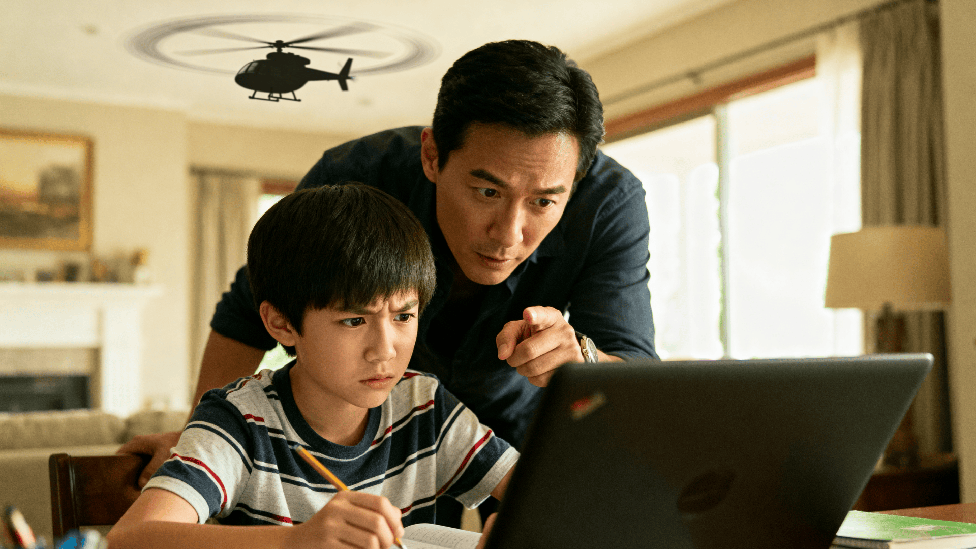 Father leans over child doing homework on laptop, pointing at screen while a toy helicopter flies overhead in a bright living room
