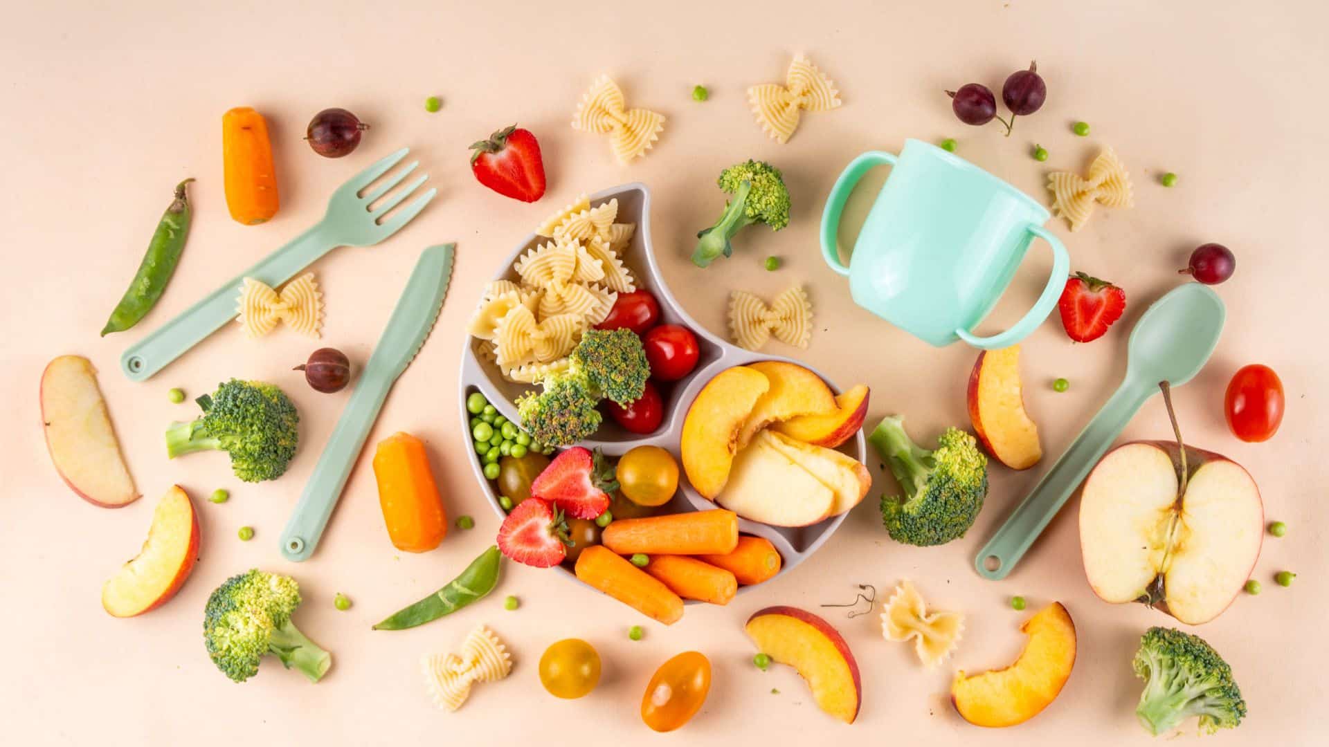 Fresh fruits, veggies, and pasta arranged with baby utensils, showing ingredients used in how to make baby food at home for healthy meals