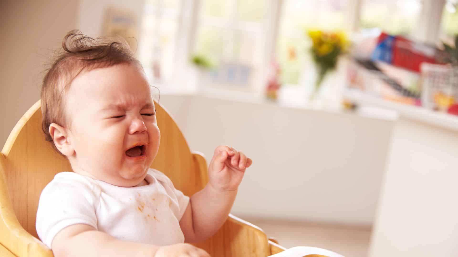 Fussy baby crying in high chair during feeding, showing discomfort, refusal to eat, and common mealtime fussiness in infants