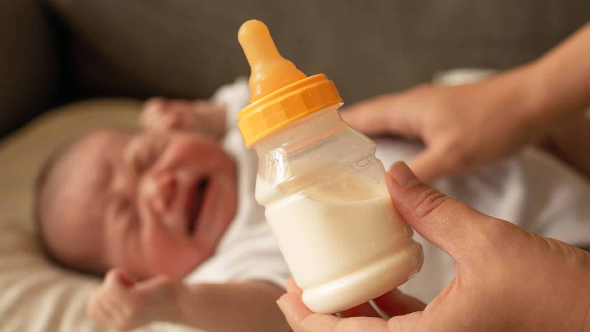Fussy baby crying while caregiver offers milk bottle, showing feeding discomfort and common soothing attempt for a restless infant