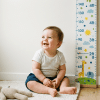 Joyful baby sitting next to a vertical growth spurt chart mounted on a light-colored wall.
