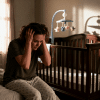 Mother experiencing postpartum rage sits on bed gripping her head in a dim nursery, surrounded by baby items, showing intense emotional overwhelm and early motherhood stress