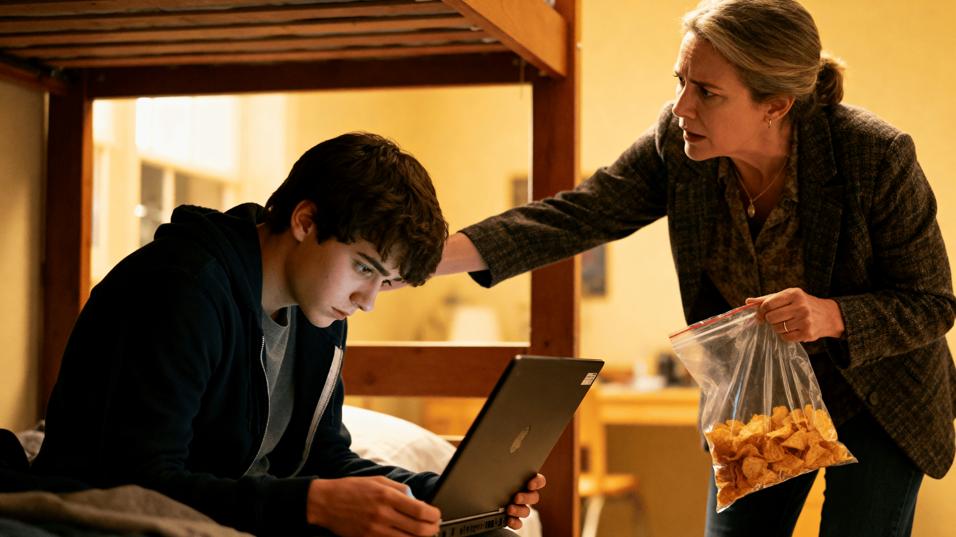 Overbearing parent hovering over teen using laptop on bed, holding snacks, monitoring activity in a tense, intrusive moment indoors.