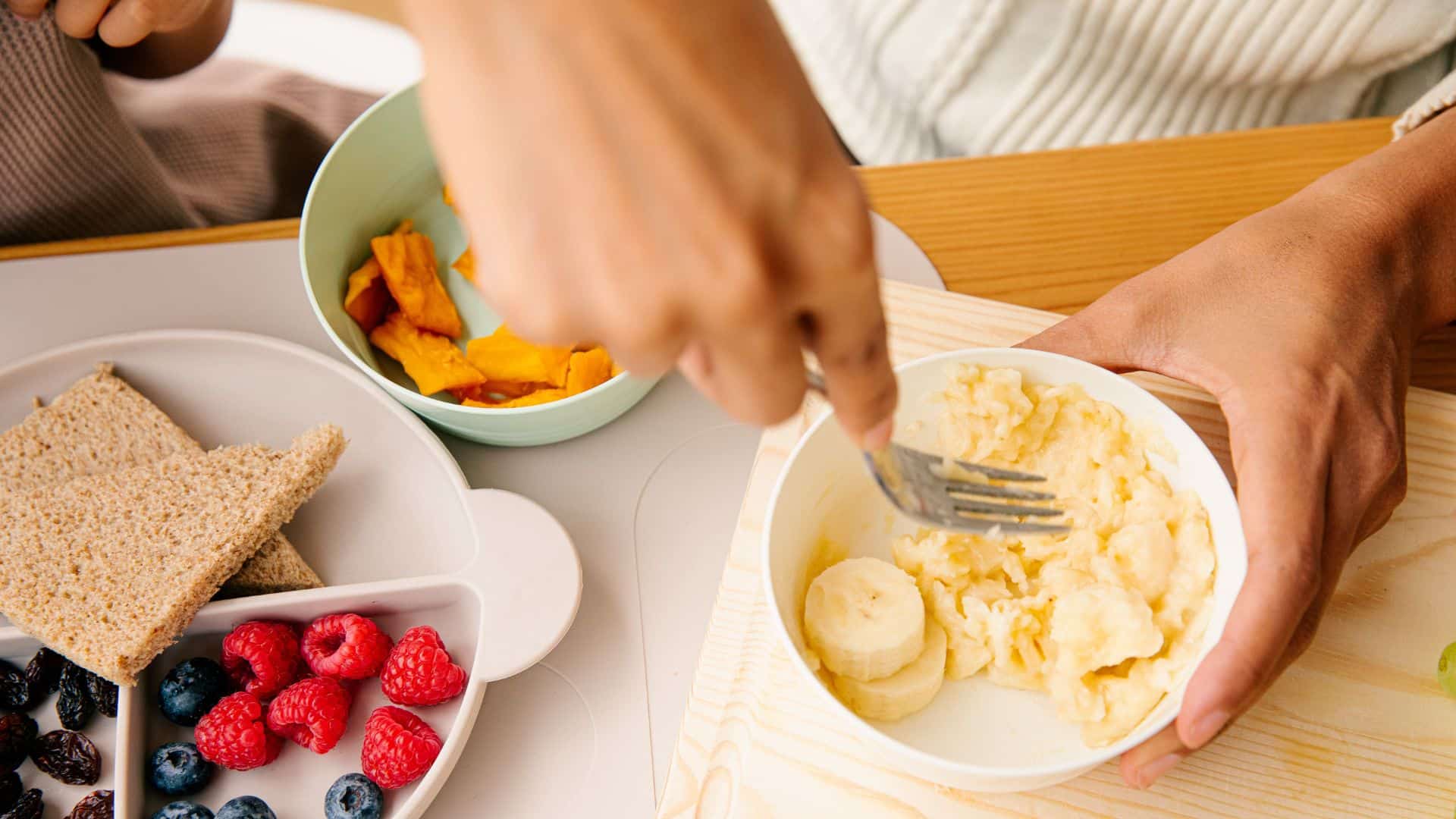 Parent mashing banana in bowl with fork beside fruits and bread, showing simple steps on how to make baby food at home for fresh healthy feeding
