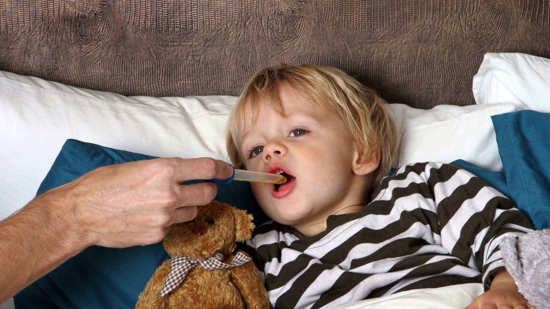 Parent using oral syringe to give medicine to toddler lying in bed, demonstrating how to get toddler to take medicine calmly and safely