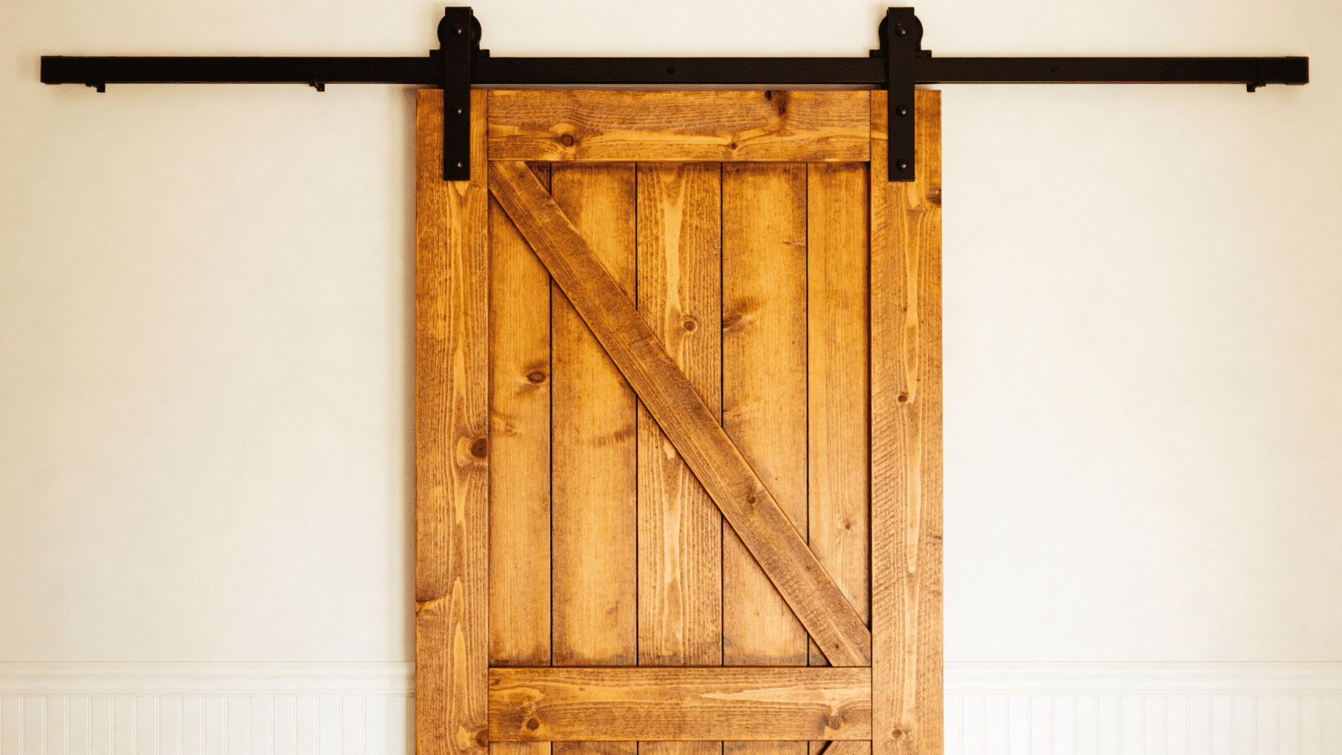 Rustic wooden barn door with diagonal brace on black sliding track, warm natural wood tones against a clean white interior wall