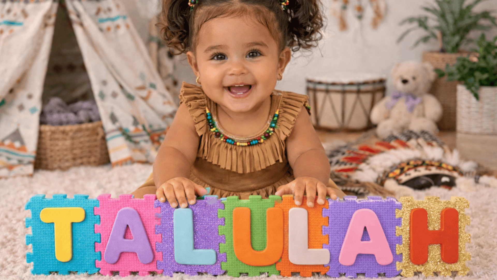 Smiling baby girl with curly pigtails sits on a carpet holding colorful foam puzzle letters spelling Tallulah in a cozy playroom.