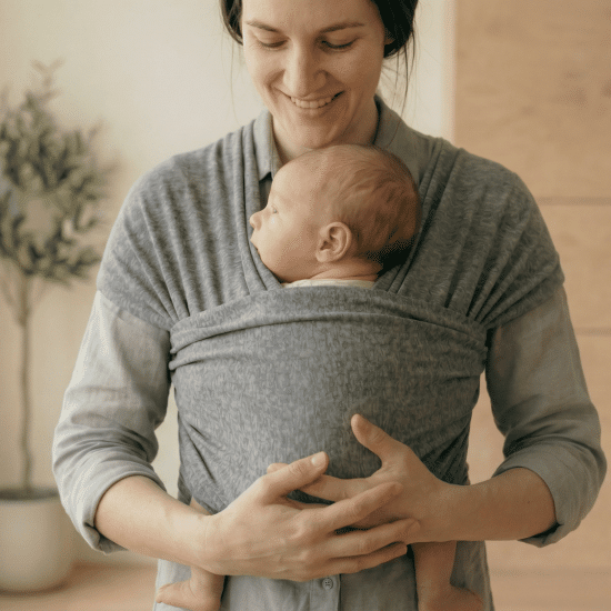 Smiling mother wearing a gray fabric wrap carrier holding a sleeping newborn baby indoors next to a potted plant.