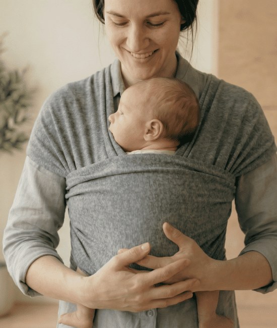 Smiling mother wearing a gray fabric wrap carrier holding a sleeping newborn baby indoors next to a potted plant.