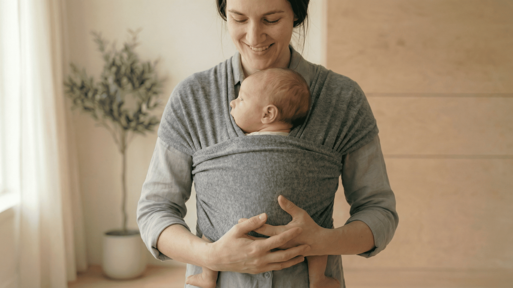 Smiling mother wearing a gray fabric wrap carrier holding a sleeping newborn baby indoors next to a potted plant.