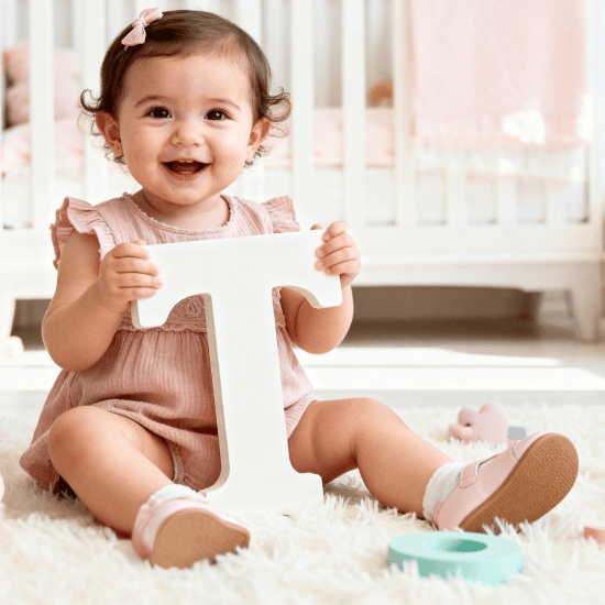 Smiling toddler girl sitting on a nursery rug holding a white letter T, surrounded by toys, blocks, and soft pastel decor.