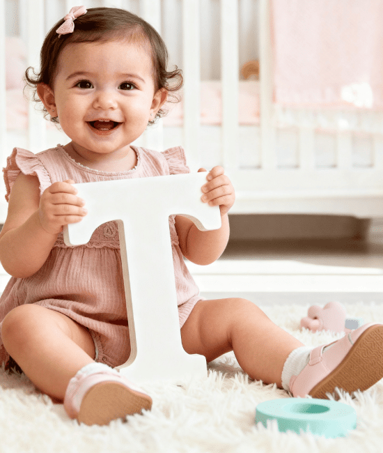 Smiling toddler girl sitting on a nursery rug holding a white letter T, surrounded by toys, blocks, and soft pastel decor.