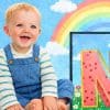 Smiling toddler in denim overalls sitting beside a large pink letter N with polka dots, rainbow and clouds background, playful scene. (1)