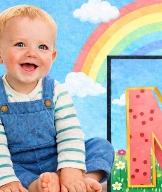 Smiling toddler in denim overalls sitting beside a large pink letter N with polka dots, rainbow and clouds background, playful scene. (1)