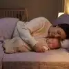 Mother and baby sleeping together in softly lit bedroom with crib in background