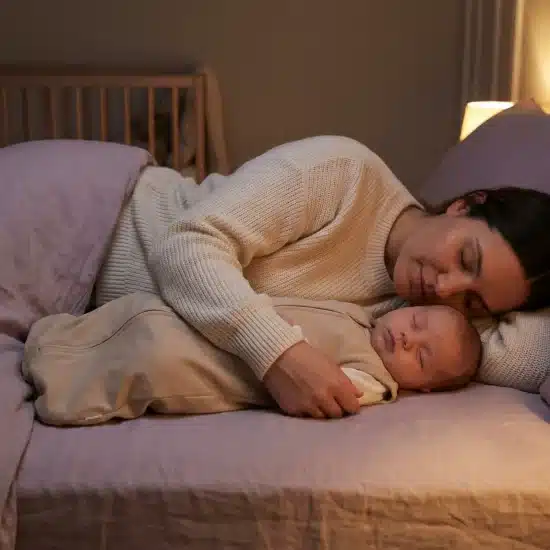 Mother and baby sleeping together in softly lit bedroom with crib in background