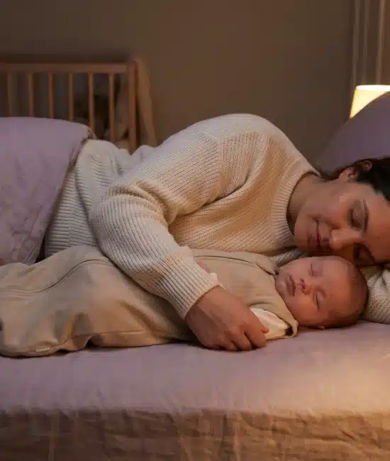 Mother and baby sleeping together in softly lit bedroom with crib in background
