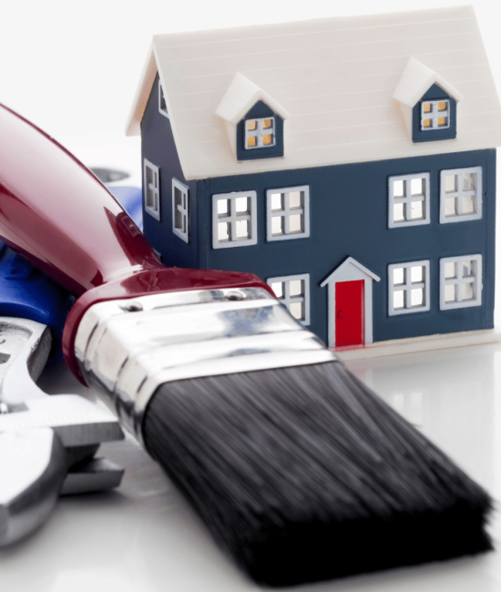 A miniature dark blue house sits behind a red-handled paintbrush and a silver adjustable wrench on a white reflective surface.