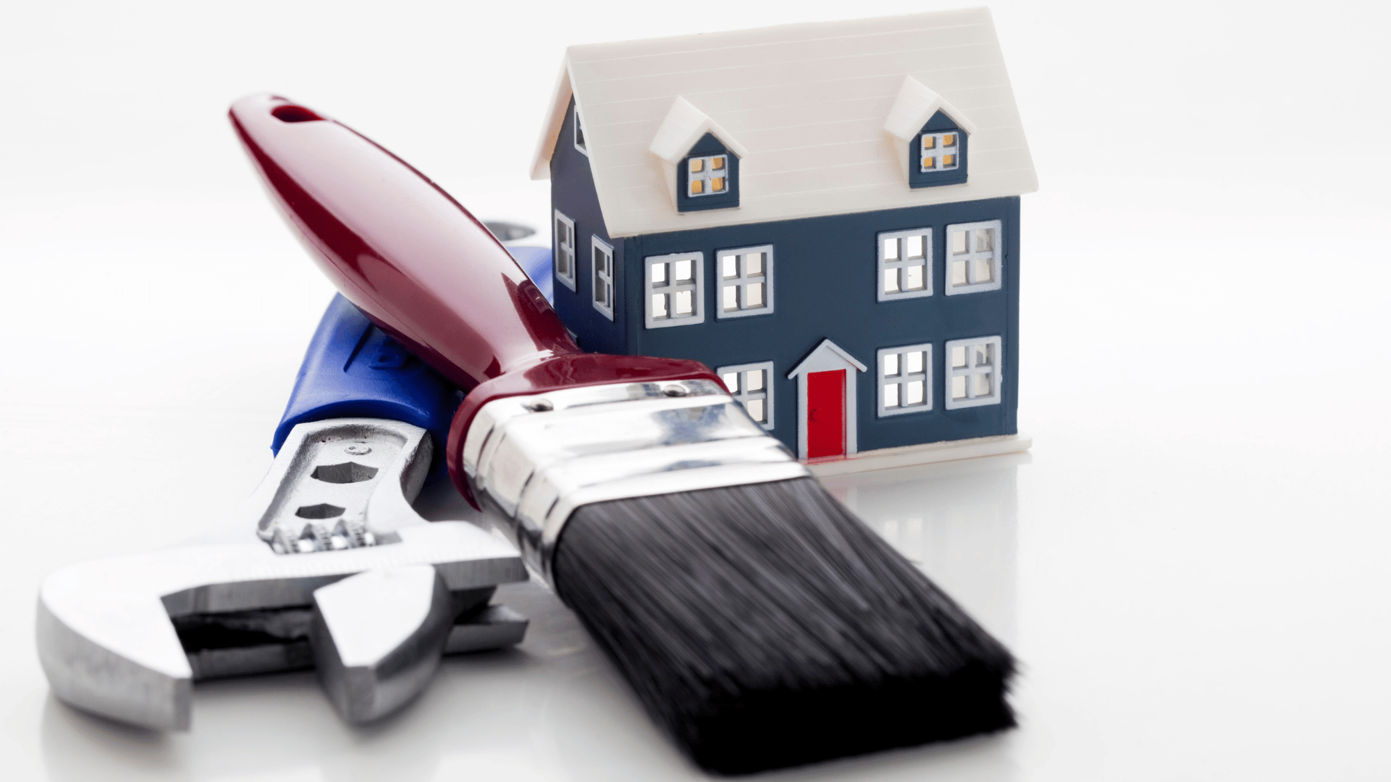 A miniature dark blue house sits behind a red-handled paintbrush and a silver adjustable wrench on a white reflective surface.