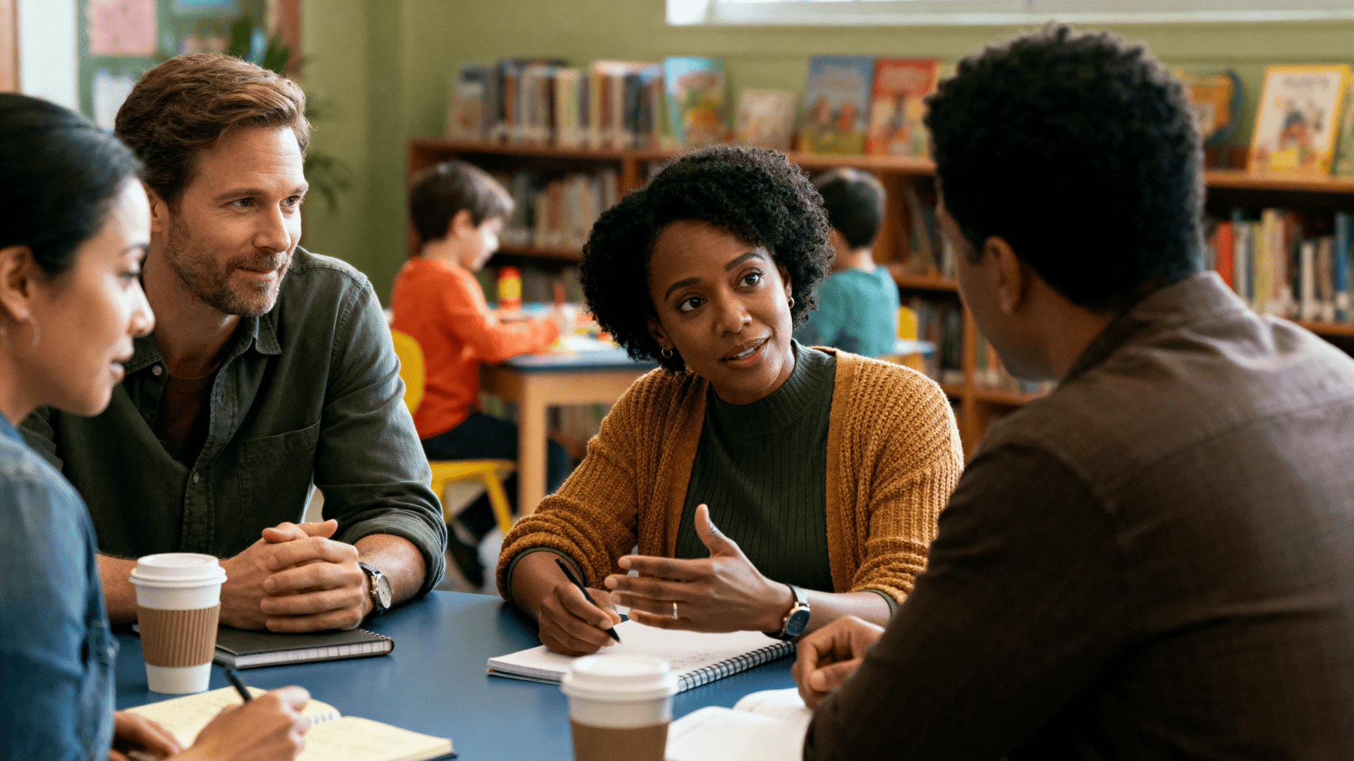Group of parents discussing homeschooling at a table with notebooks and coffee, in a warm community space with kids in background.