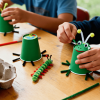 Kids making bug crafts with green paper cups, pipe cleaners, and googly eyes at a table with paint, scissors, and egg cartons
