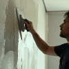 Man applying drywall texture with a trowel on an interior wall, creating patterned finish in a softly lit modern room under renovation
