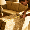 Man measuring sofa width with tape in warm living room, vintage couch, focused posture, soft lighting, home interior scene