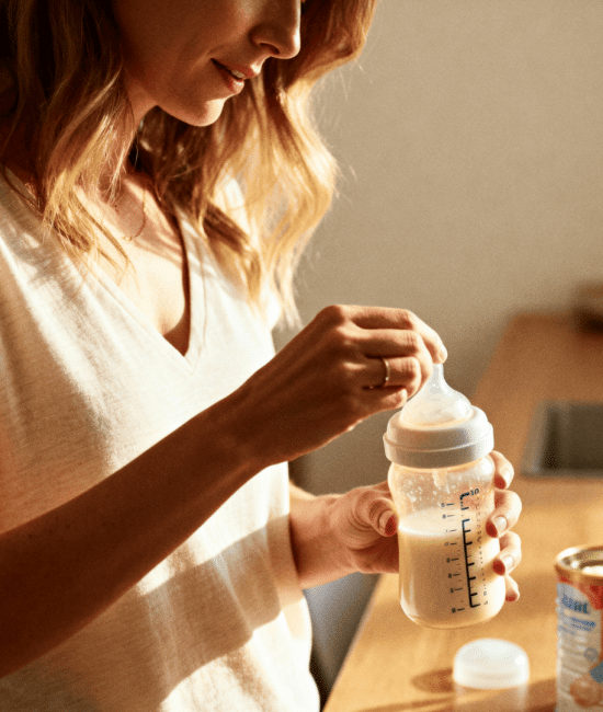 Mother mixing baby formula in a bottle at home kitchen, warm natural light, preparing infant feeding with calm nurturing atmosphere