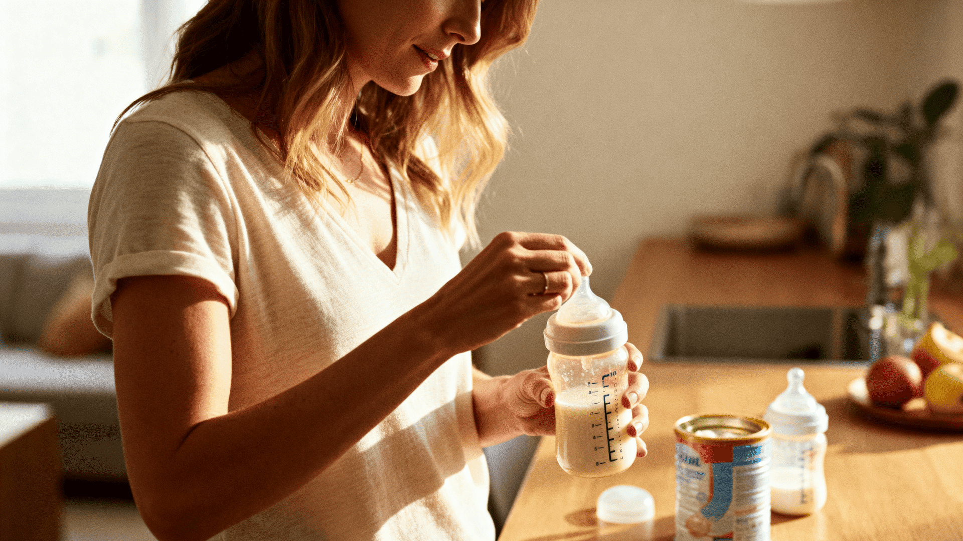 Mother mixing baby formula in a bottle at home kitchen, warm natural light, preparing infant feeding with calm nurturing atmosphere