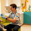 Smiling pediatric dentist examines a calm baby on a parent’s lap in a colorful, child-friendly clinic with toys and bright decor