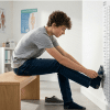 Teen boy on a bench in a bright clinic, measuring his socked foot against a size chart on a wall.