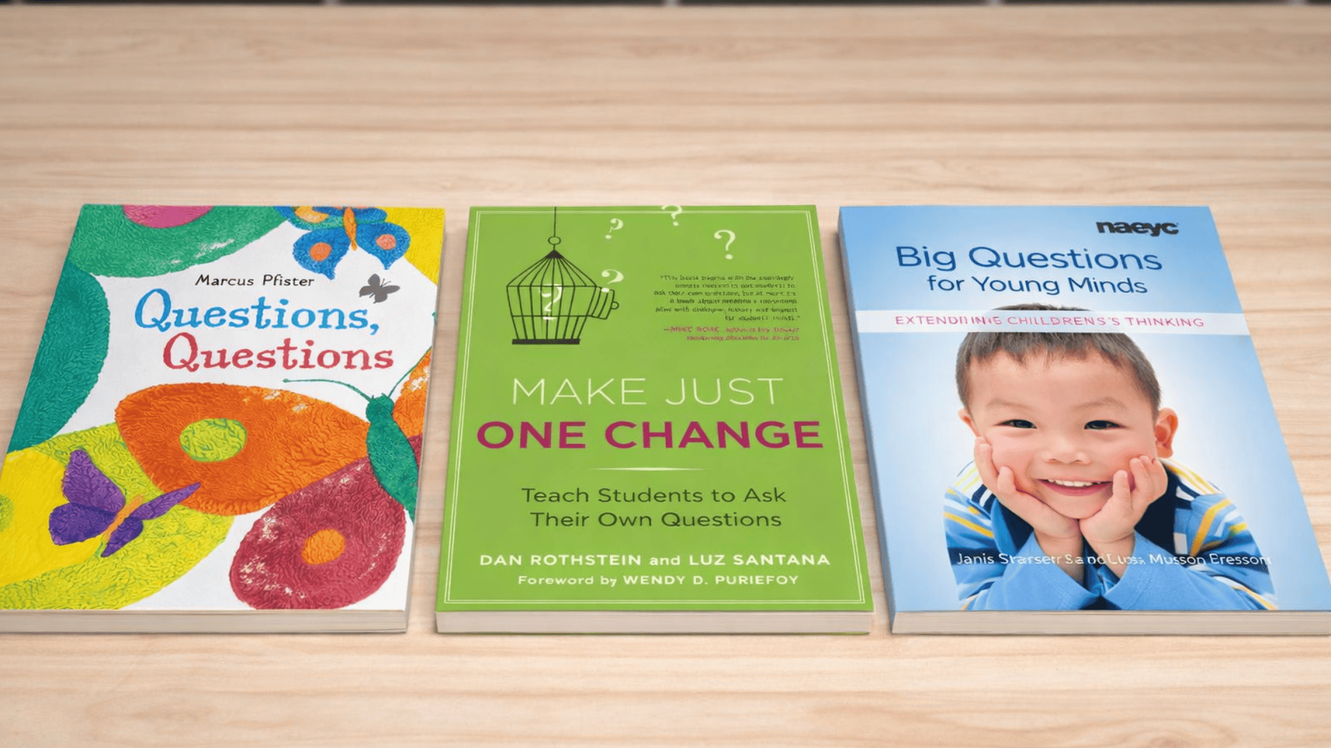 Three books lie flat on a wooden table “Questions, Questions,” “Make Just One Change,” and “Big Questions for Young Minds.”