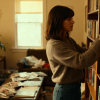 Woman organizing books on a wooden shelf in cozy living room, warm lighting, coffee table cluttered with magazines, home interior scene