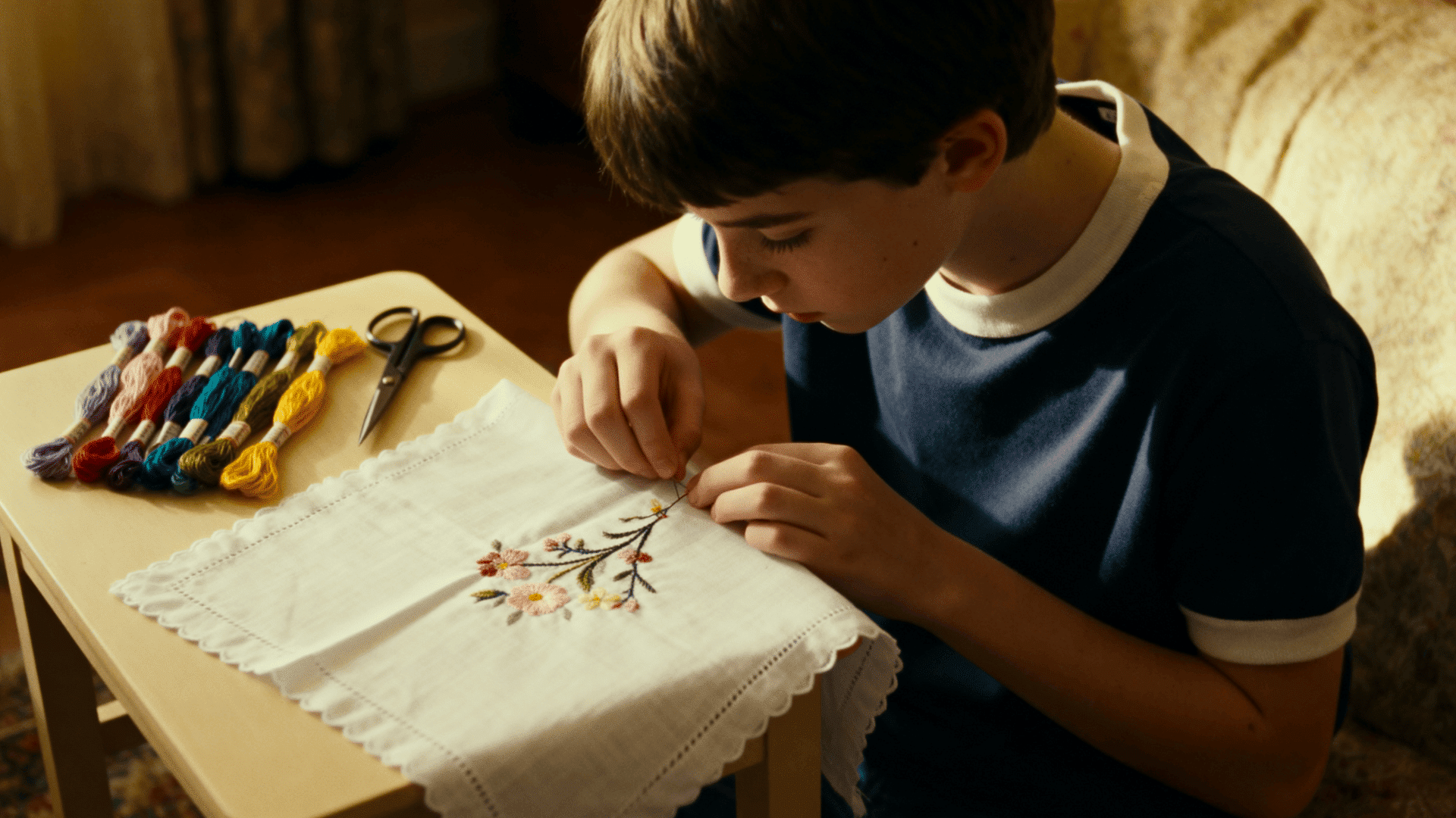 Young boy embroidering floral pattern on white cloth at table, colorful threads and scissors nearby, focused crafting scene indoors