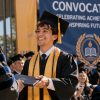 A smiling young man in a graduation cap and gown receives a diploma during a convocation ceremony with an audience clapping.