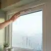 Hand adjusting a ventilation device on a window with visible condensation and indoor potted plant nearby