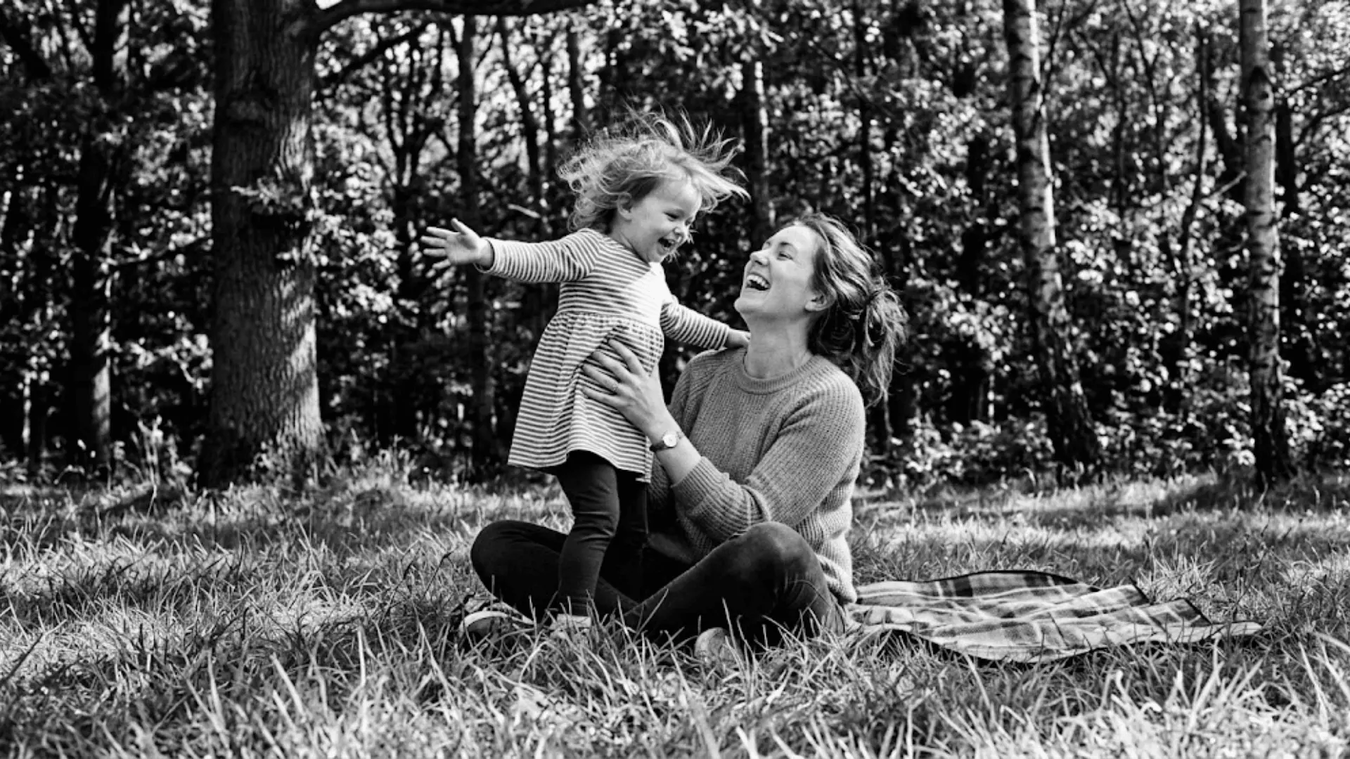 black and white candid of a laughing mother balancing her toddler on her knees on a sunny grass lawn