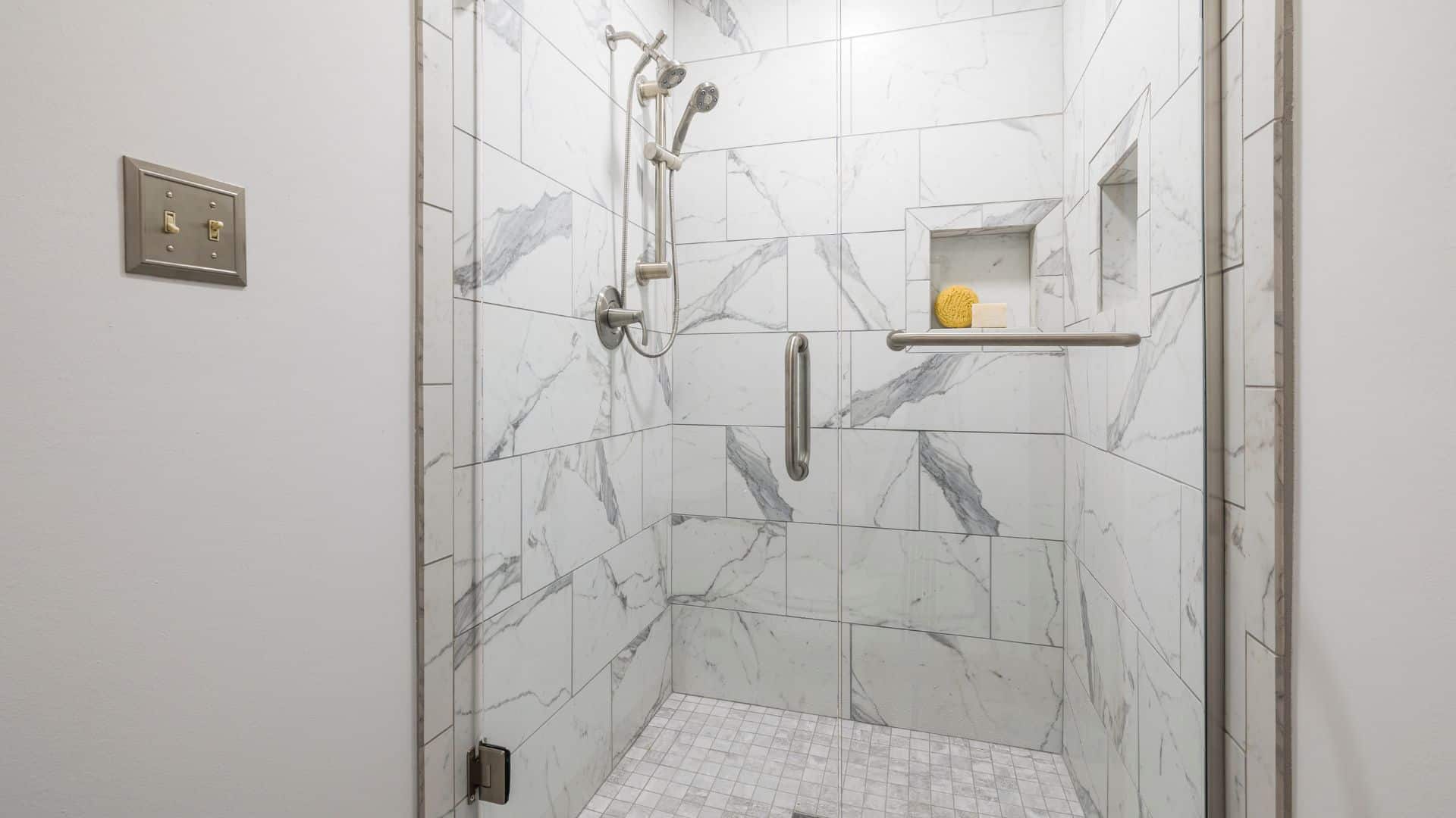 Clean white marble shower with glass door, chrome fixtures, built-in shelf, and small mosaic tile floor