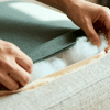 Close-up of hands inserting fresh polyfill into a couch cushion through a zipper opening, showing fabric texture and cushion repair process