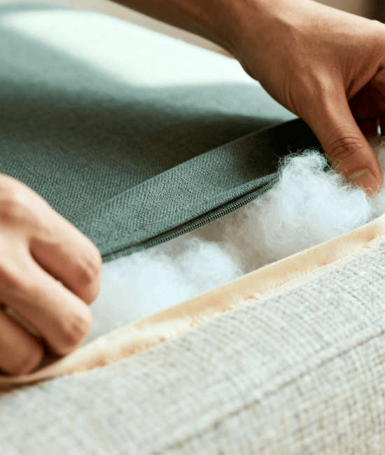 Close-up of hands inserting fresh polyfill into a couch cushion through a zipper opening, showing fabric texture and cushion repair process