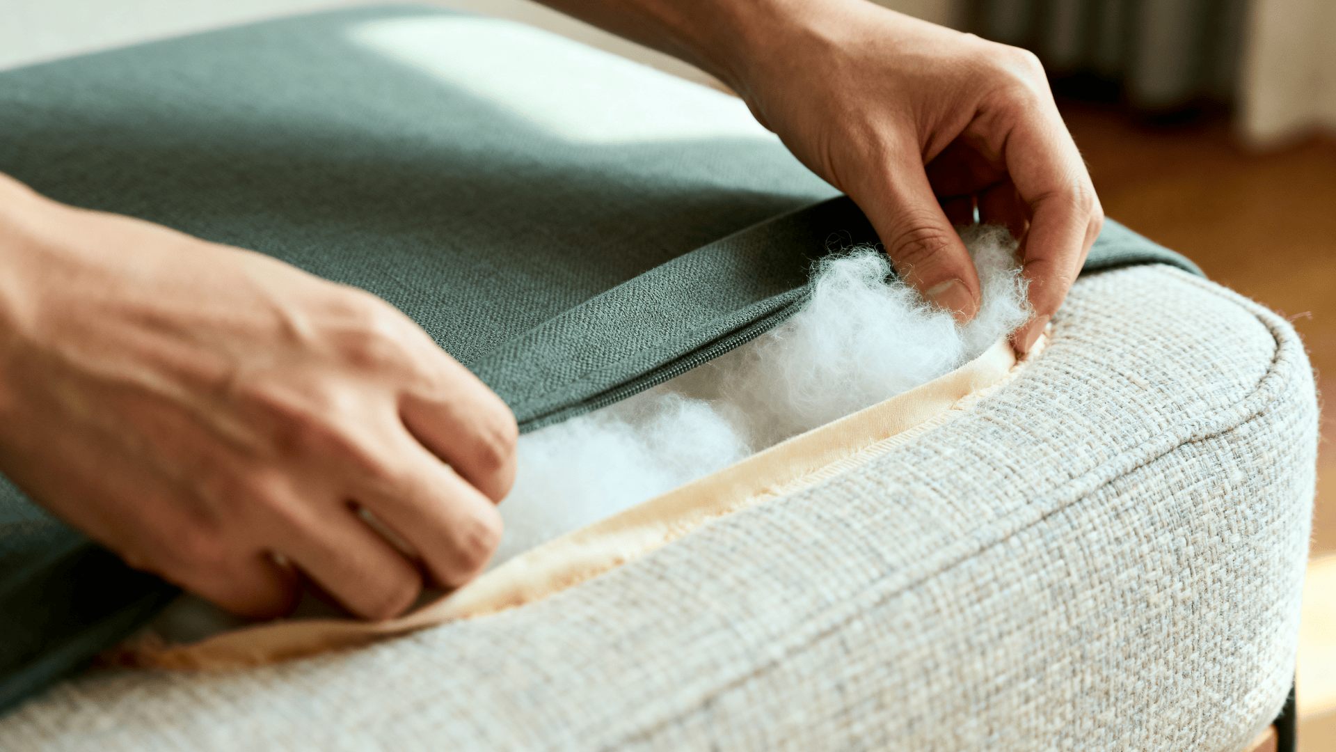 Close-up of hands inserting fresh polyfill into a couch cushion through a zipper opening, showing fabric texture and cushion repair process